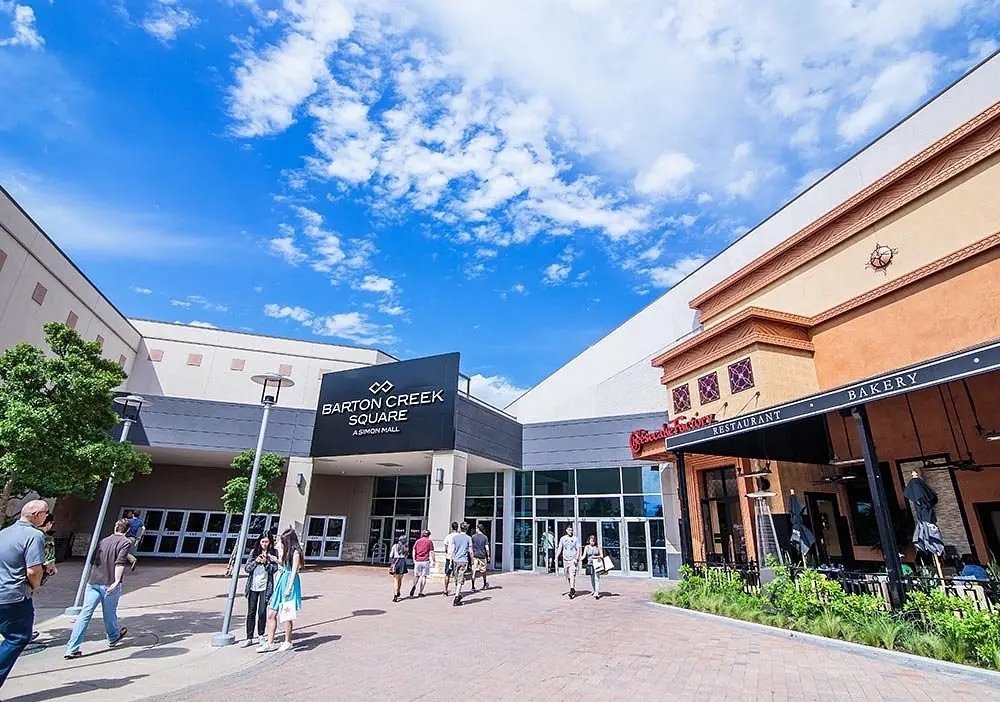 People walking in front of Barton Creek Square shopping center under a clear blue sky.