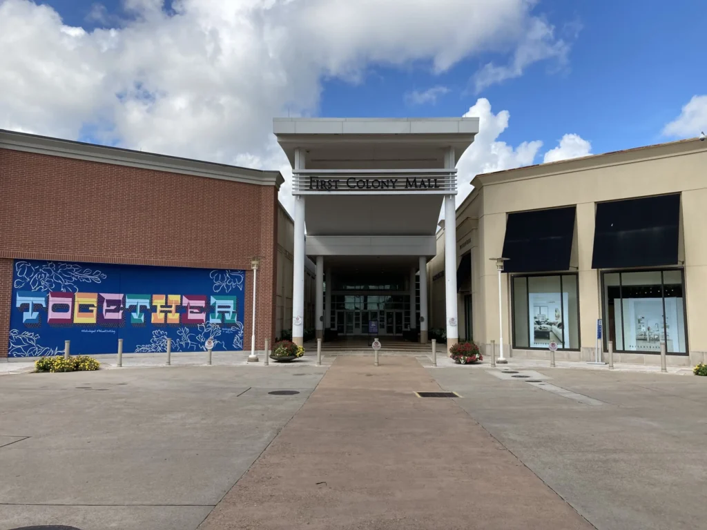 Exterior of First Colony Mall with palm trees and people walking around.