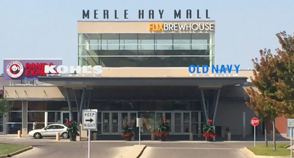 Shopping center exterior with 'Merle Hay Mall' sign and parking lot filled with cars.