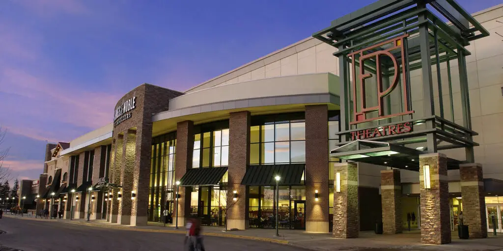 A bustling shopping center at dusk, its front entrance illuminated by warm lights, beckoning visitors with promises of retail therapy and excitement.