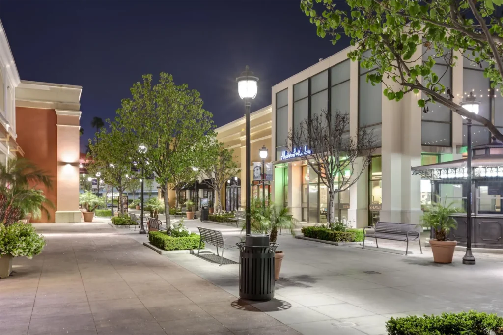 A night view of a shopping center with brightly lit storefronts and bustling activity under the dark sky.