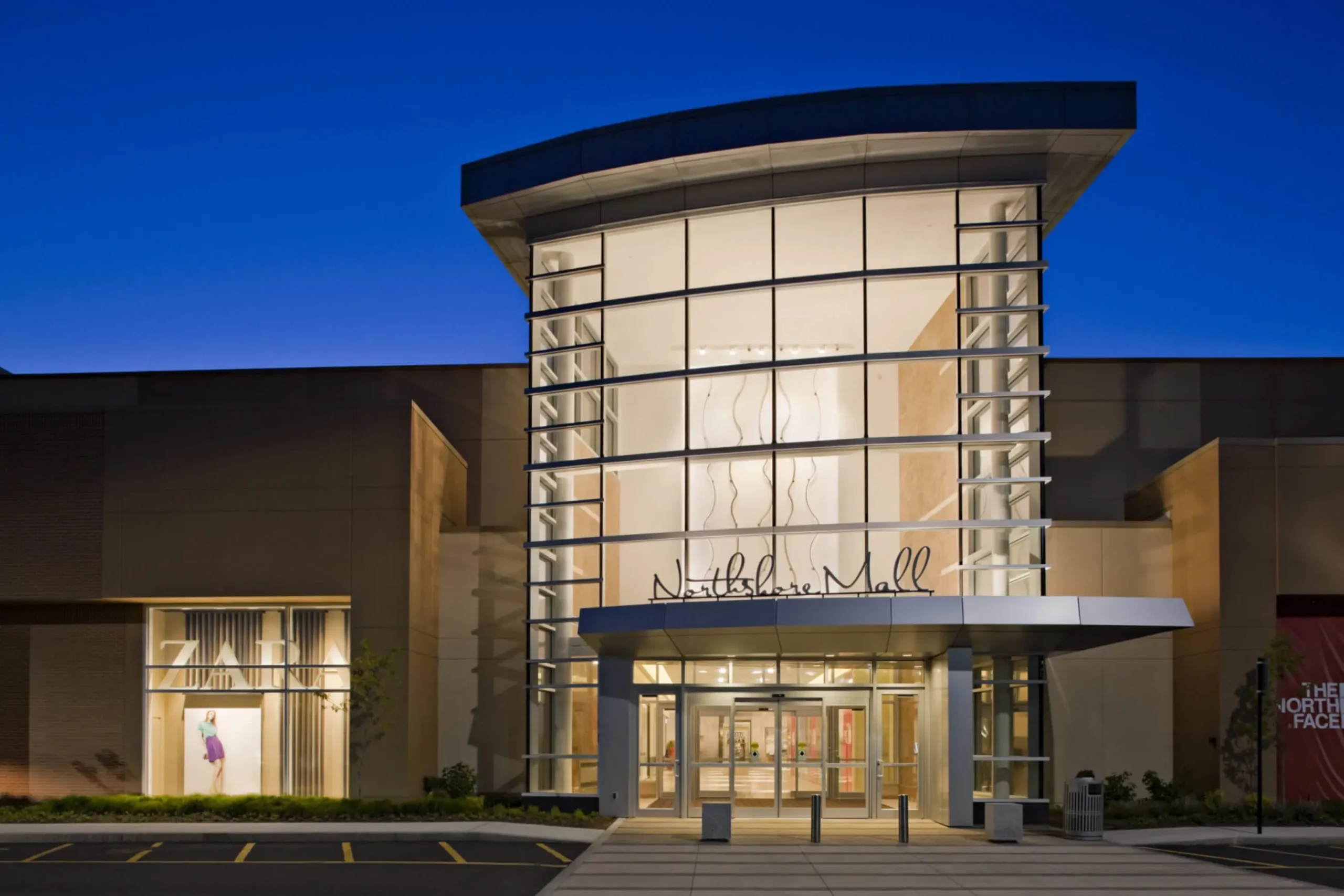 Entrance to a bustling shopping center at night, with bright lights illuminating the facade and people entering and exiting.