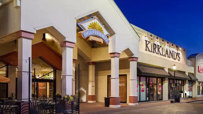 The front entrance of a shopping center at dusk: A well-lit entrance with people entering and exiting the building under a colorful sky.