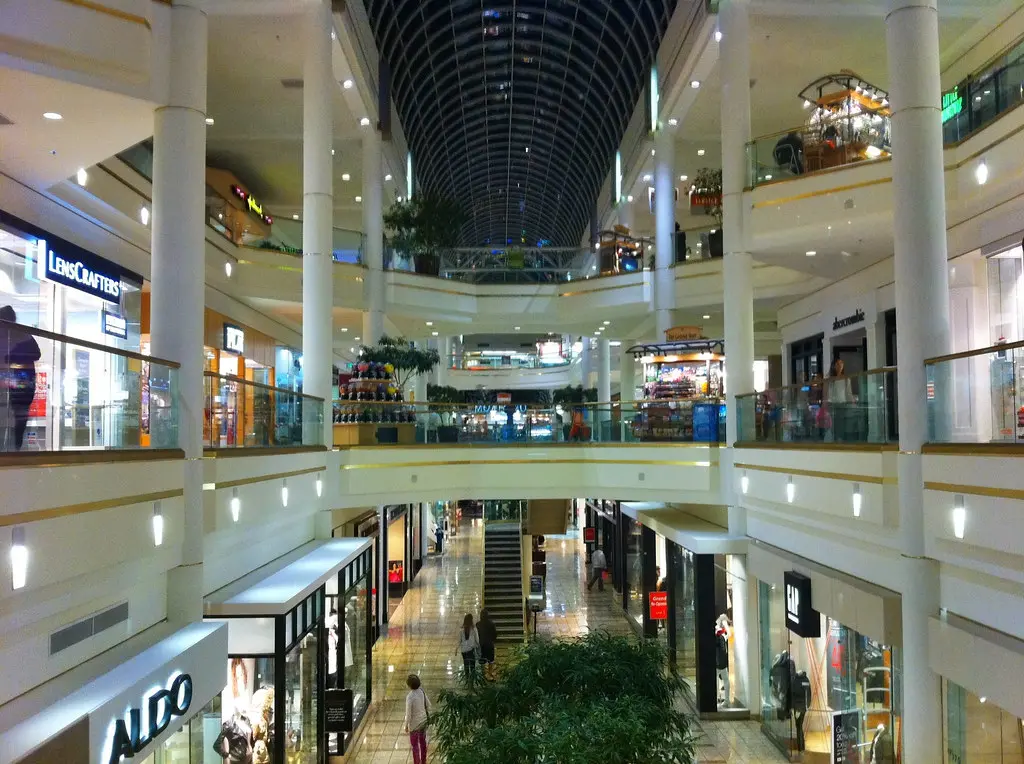 A bustling shopping mall with numerous stores, escalators, and shoppers browsing through various products.