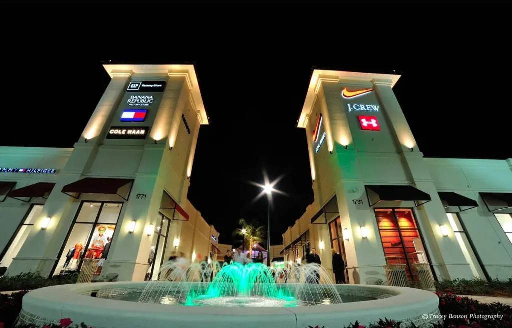 Illuminated fountain in front of Tanger Outlets Palm Beach Mall building at night.